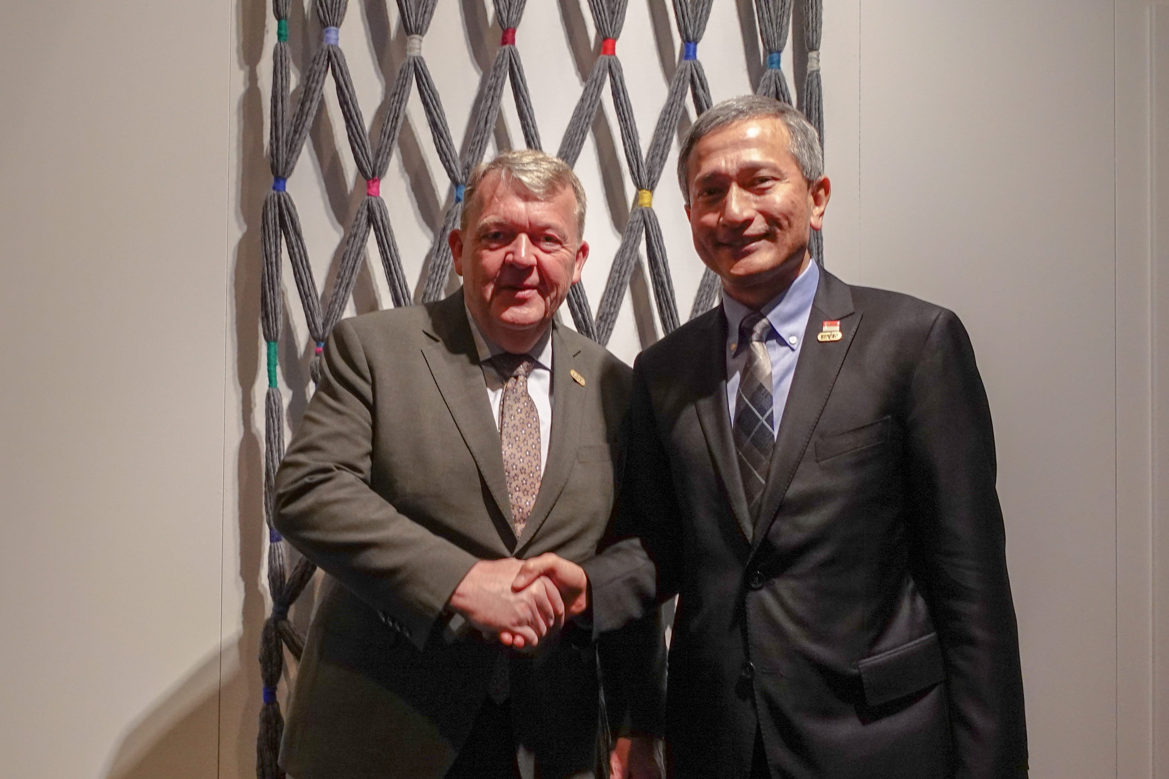 Two men in suits shake hands, posed in front of a gray fiber art wall hanging.
